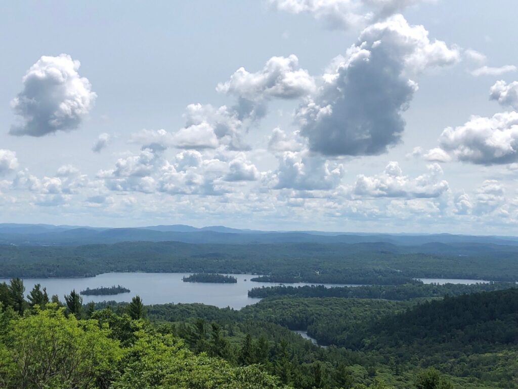 Vue du haut de la tour Mont Morisette, offrant une vue élevée au-dessus de la forêt, avec une étendue d'eau au loin, des collines en arrière-plan et un ciel bleu avec quelques gros nuages blancs.